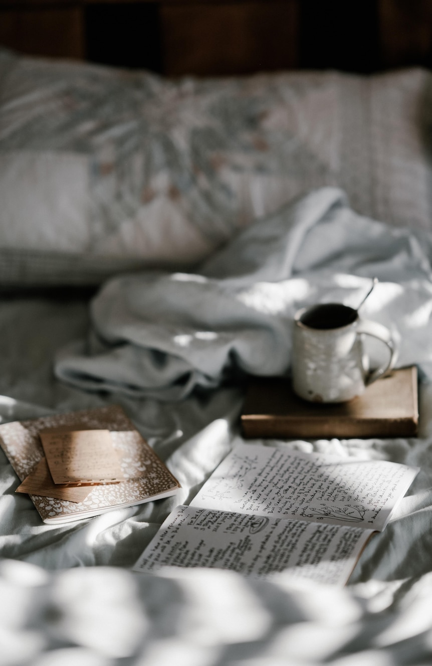 Coffee and notebooks and open journal on bed with white bed linen and dappled morning light.
