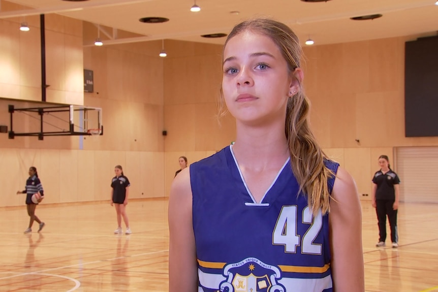 A young girl with blonde hair in a ponytail wearing a blue basketball top stands on a basketball court with players behind her