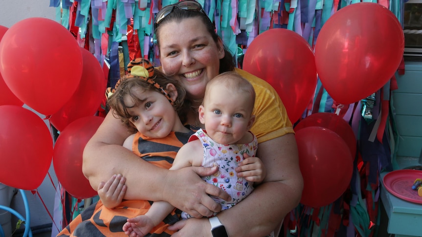 A mother hugs her two daughters with red balloons on either side.