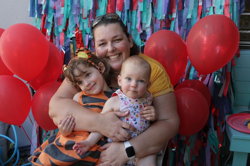A mother hugs her two daughters with red balloons on either side.