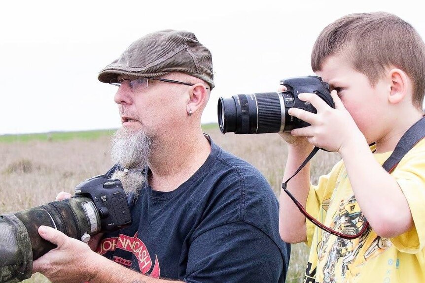 Grey-bearded man wearing a flat cap with a camera resting in his hands and a boy in yellow T-shirt with camera raised