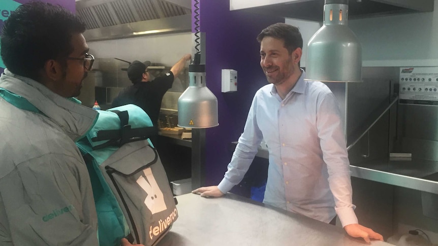 The general manager of Deliveroo Levi Aron stands in a kitchen in Melbourne a delivery driver collects a bag of food.