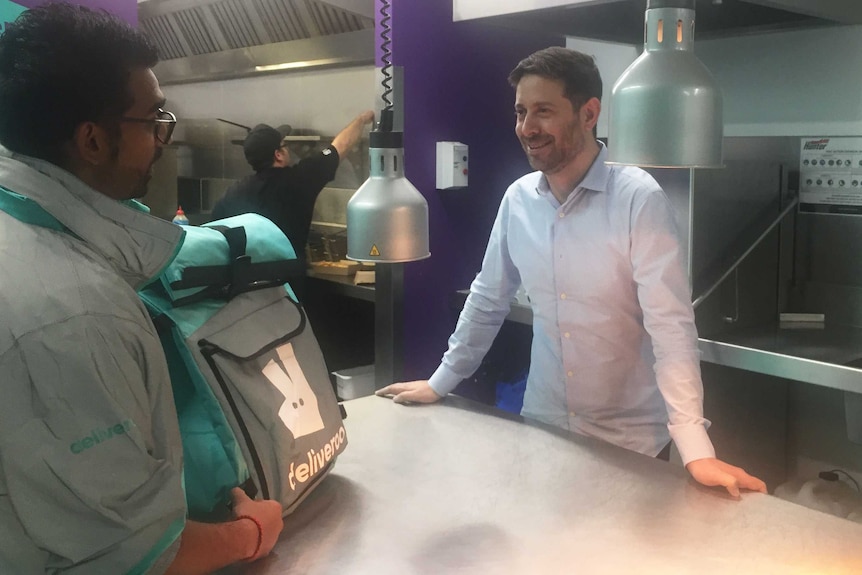 The general manager of Deliveroo Levi Aron stands in a kitchen in Melbourne a delivery driver collects a bag of food.