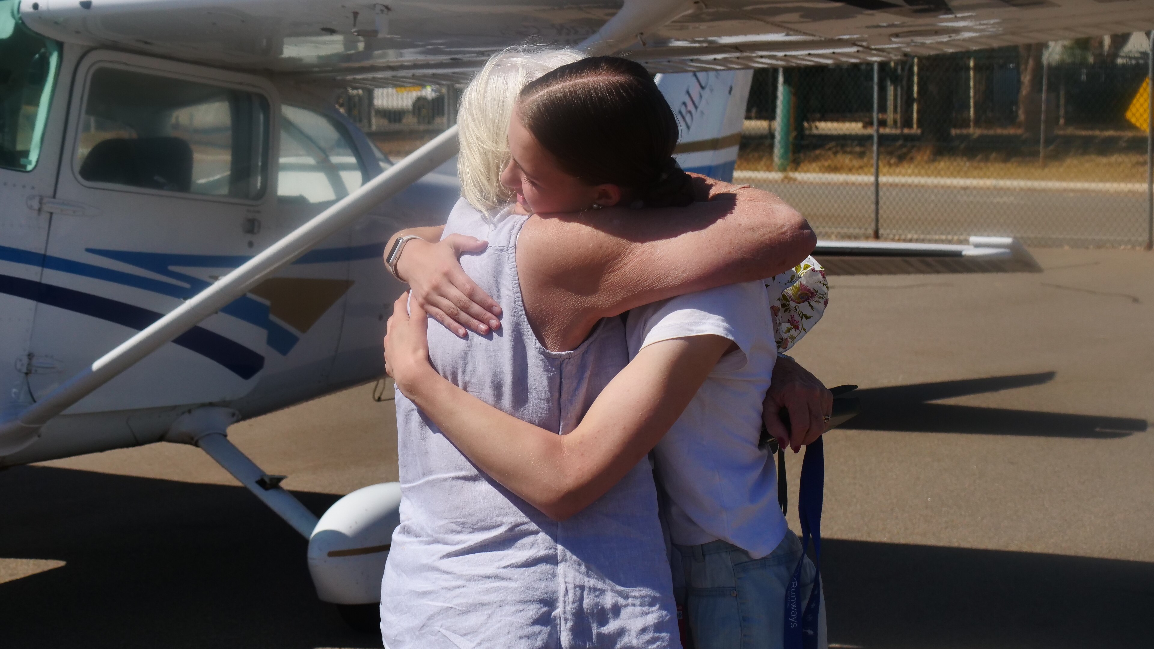A young woman with brown hair and an older woman with white hair hug in the sun, in front of a small airplane.