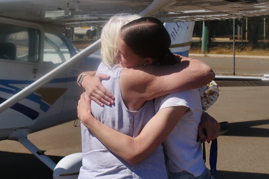 A young woman with brown hair and an older woman with white hair hug in the sun, in front of a small airplane.
