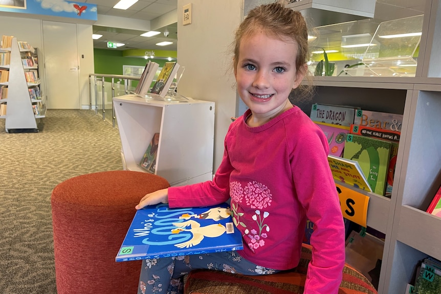A young girls sits in a library holding a book and smiling.