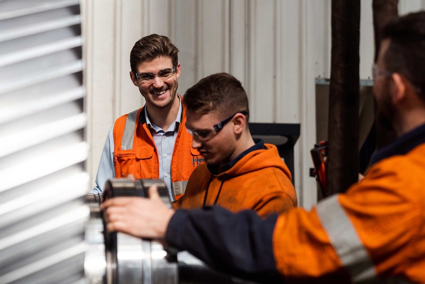 A man in hi-vis clothing and safety glasses smiles and laughs talking to two workers in a workshop.
