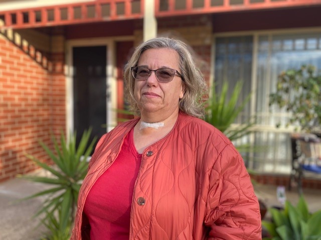 A middle-aged woman with a peach-coloured shirt stands in front of a red brick home.