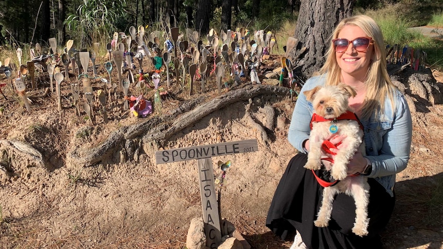 a young woman smiles at the camera holding her small dog. Behind her are hundreds of wooden spoons with smiley faces in the park