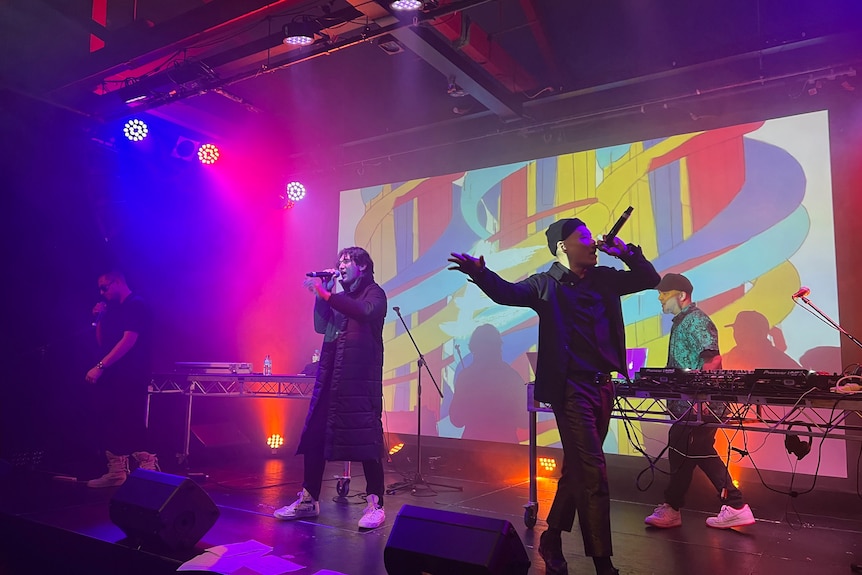 Four Asian Australian musicians, including three vocalists, perform on stage, in front of a screen with bright visuals