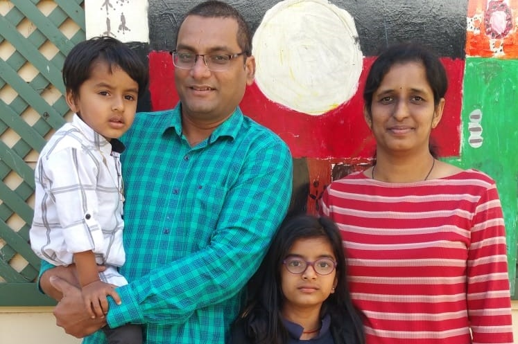 Family standing in front of Indigenous artwork, man holding son on left, eight-year-old girl in middle and wife on right