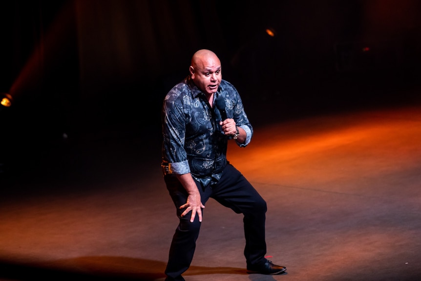 A 50-something Aboriginal man squats down on stage, one hand on his thigh and the other holding a microphone to his face