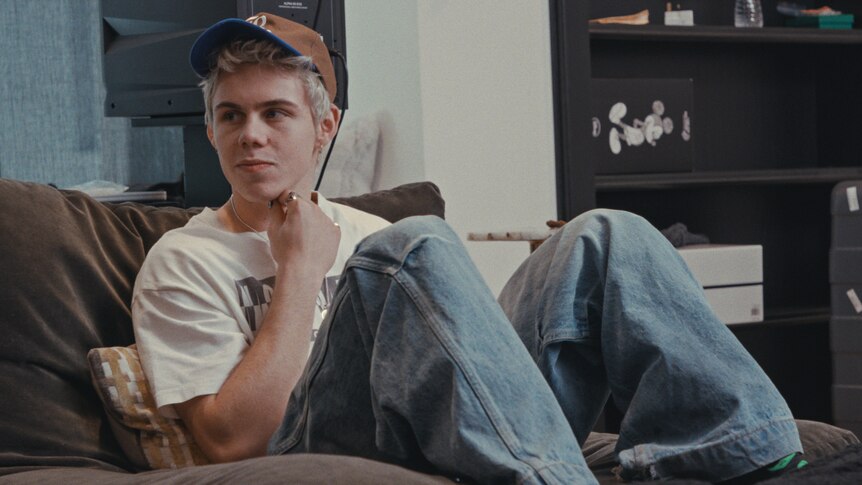 The Kid Laroi sits with his feet up on a couch while wearing baggy jeans, a white shirt and a brown cap