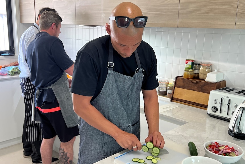 A man cuts up cucumber on a kitchen bench as others work behind him in the kitchen