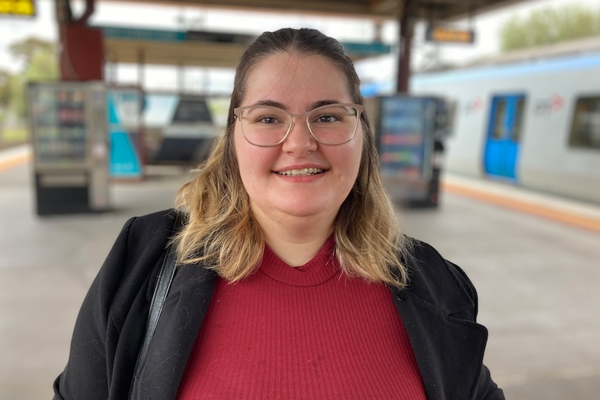 Brittany smiles as she stands at a train platform, in a jacket and red jumper.