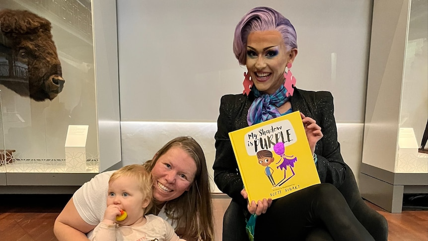 Drag Queen sitting on a chair next to woman with toddler. Drag Queen is holding a book titled "My Shadow Is Purple"