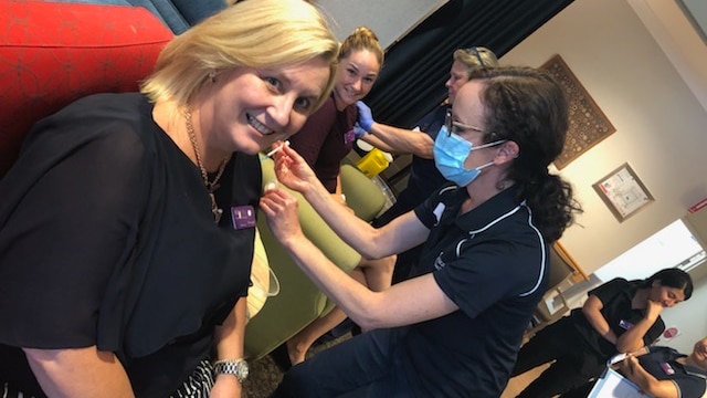 Two women sit in chairs while two nurses deliver a vaccination jab.