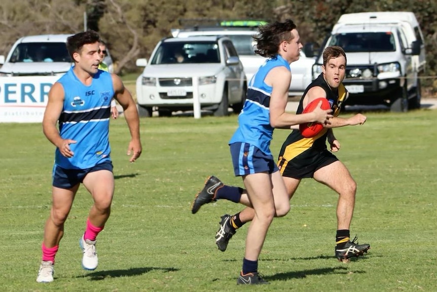 An Australian rules football player wearing light blue holds a football as a teammmate and another player trail