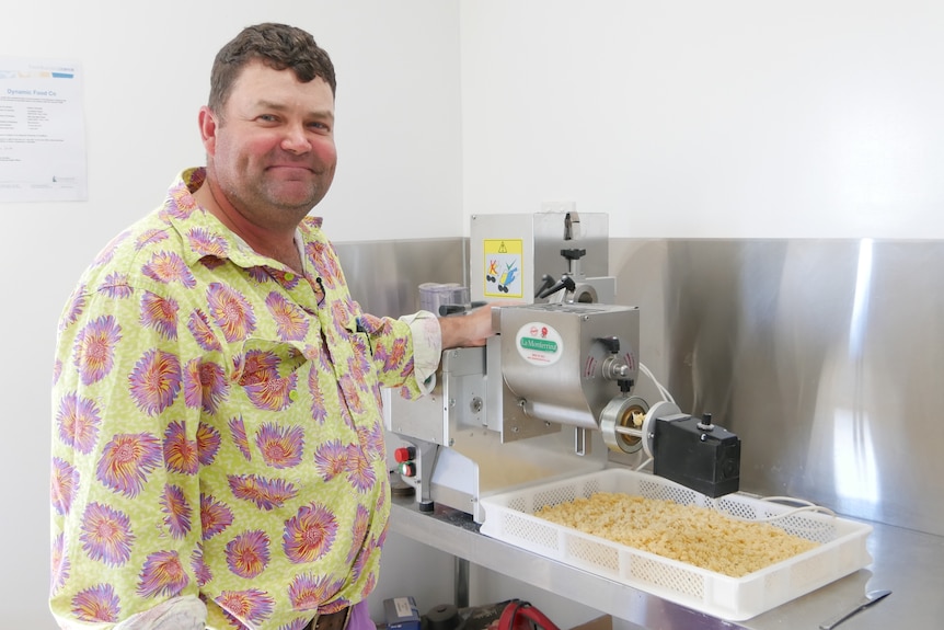 A man in a lime and pink floral work shirt smiles. He is standing next to a pasta machine