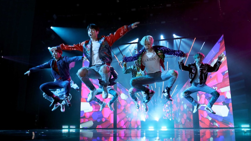 The seven members of BTS jump in unison on a backlit stage.
