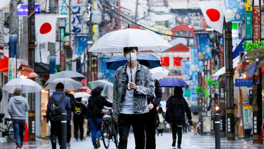 A Japanese man in a face mask holds an umbrella while walking down a Tokyo street