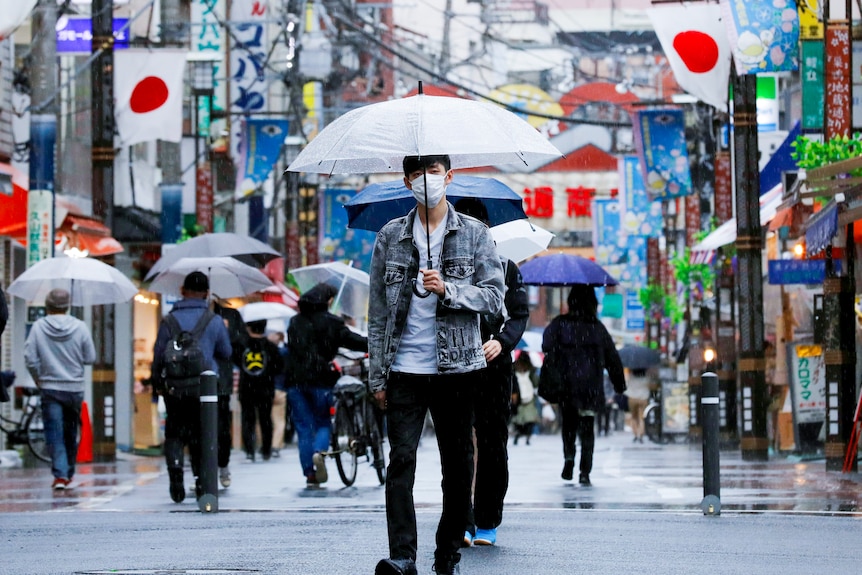 A Japanese man in a face mask holds an umbrella while walking down a Tokyo street