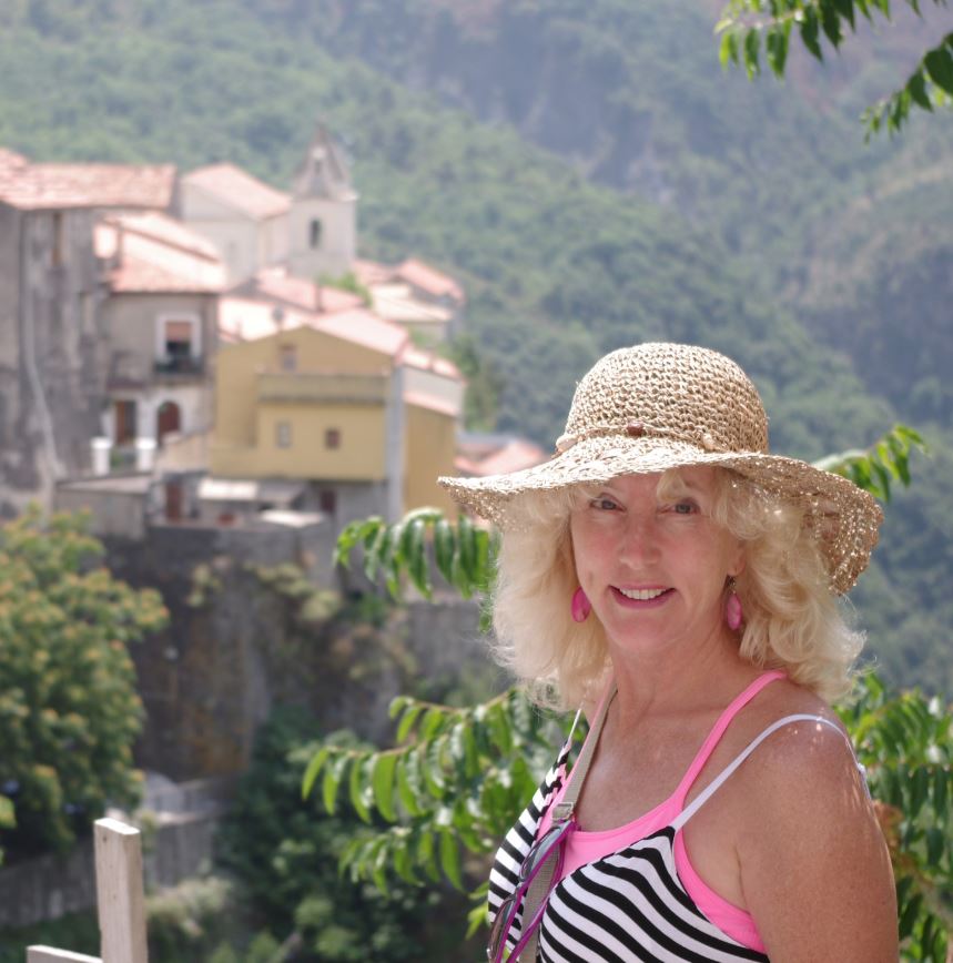 A woman smiles while wearing a hat and posing in front of a mountainous landscape.