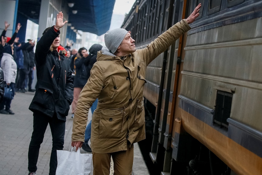 Men stand on a train station platform, waving goodbye to people on an evacuation train.