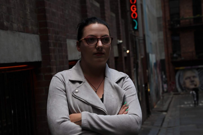 Ash looks at the camera from a city laneway, with a bar sign behind her and graffiti at the end of the lane.