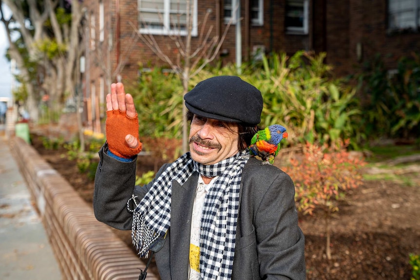 Mr Gawronski sits by the side of his apartment block with a parrot on his shoulder waving at a passer-by.