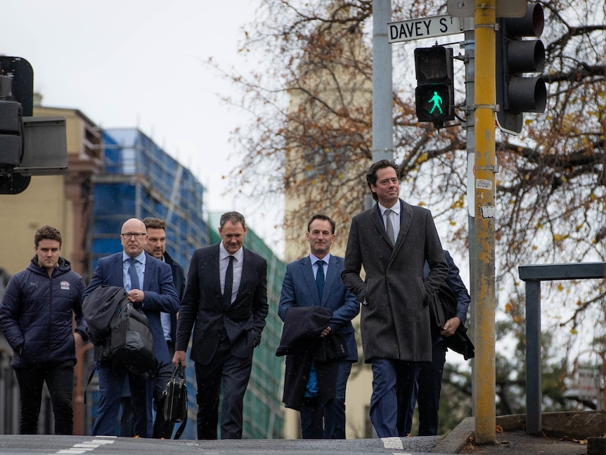 Seven men in suits cross a Hobart street at a pedestrian crossing.