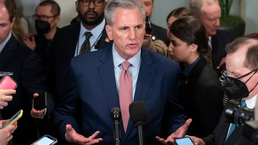 A man in a suit speaks surrounded by a media scrum.