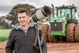 Farmer Drew Chislett standing in a paddock with a tractor in the background with his hefty 400mm lens slung over his shoulder.