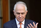 Malcolm Turnbull frowns, looks down and raises his open hand. He is standing at a microphone.