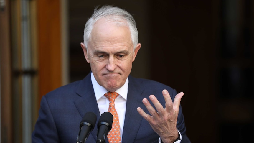 Malcolm Turnbull frowns, looks down and raises his open hand. He is standing at a microphone.