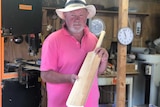 A man standing in a shed holds up a hand-carved cricket bat.