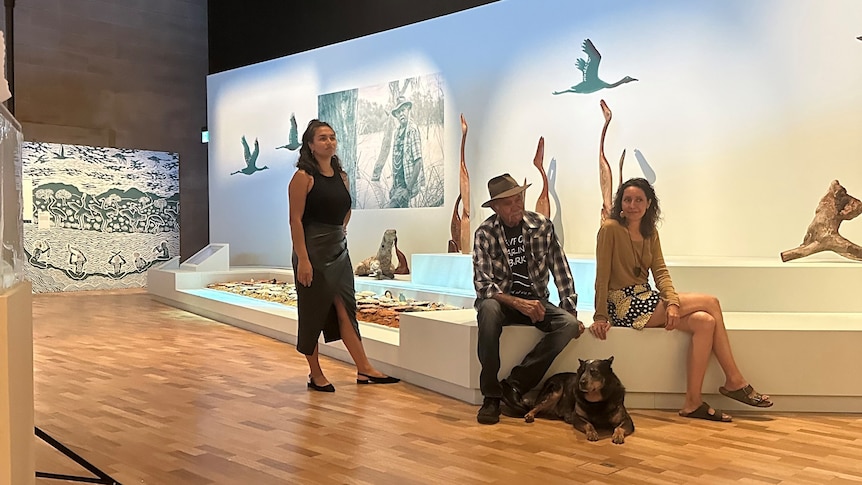 A woman standing next to a man, dog and woman sitting on a bench in front of a series of artworks