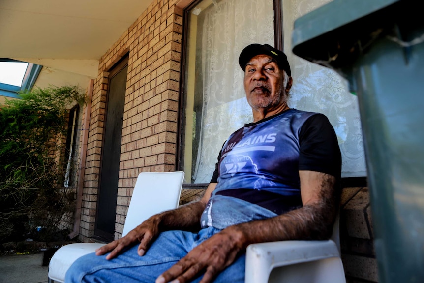 Man wearing cap and jeans sits on white plastic chair next to green bin outside brick home exterior