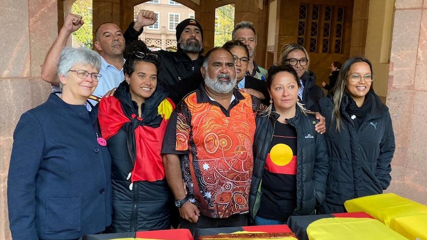 A group of male and female Indigenous Australians stand together inside sandstone building.