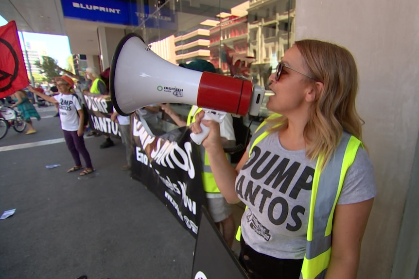 A woman with blonde hair wearing a shirt saying DUMP SANTOS speaks into a bullhorn