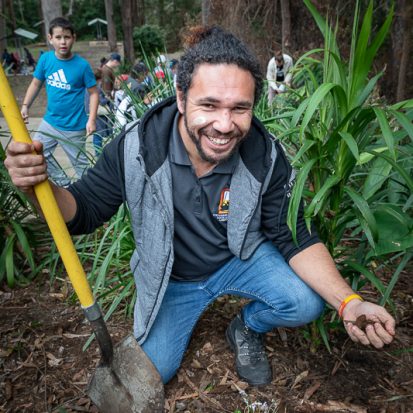 Nathan Brennan smiling, kneeling in front of some plants, and holding a shovel.