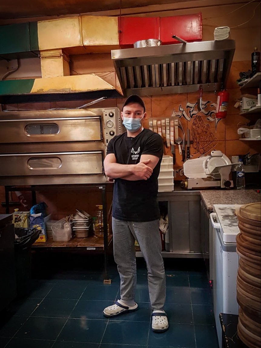 A man in a blue face mask stands with his arms folded in a pizzeria kitchen