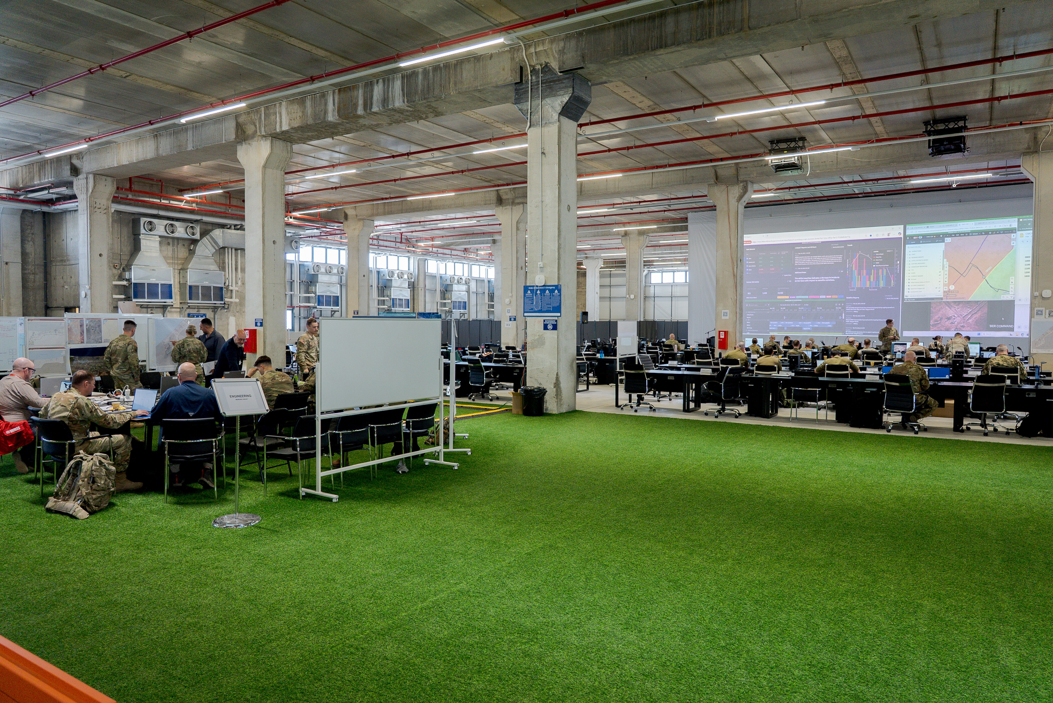 A large warehouse with industrial columns and fake grass floor full of screens, desks and people, some in military uniforms.