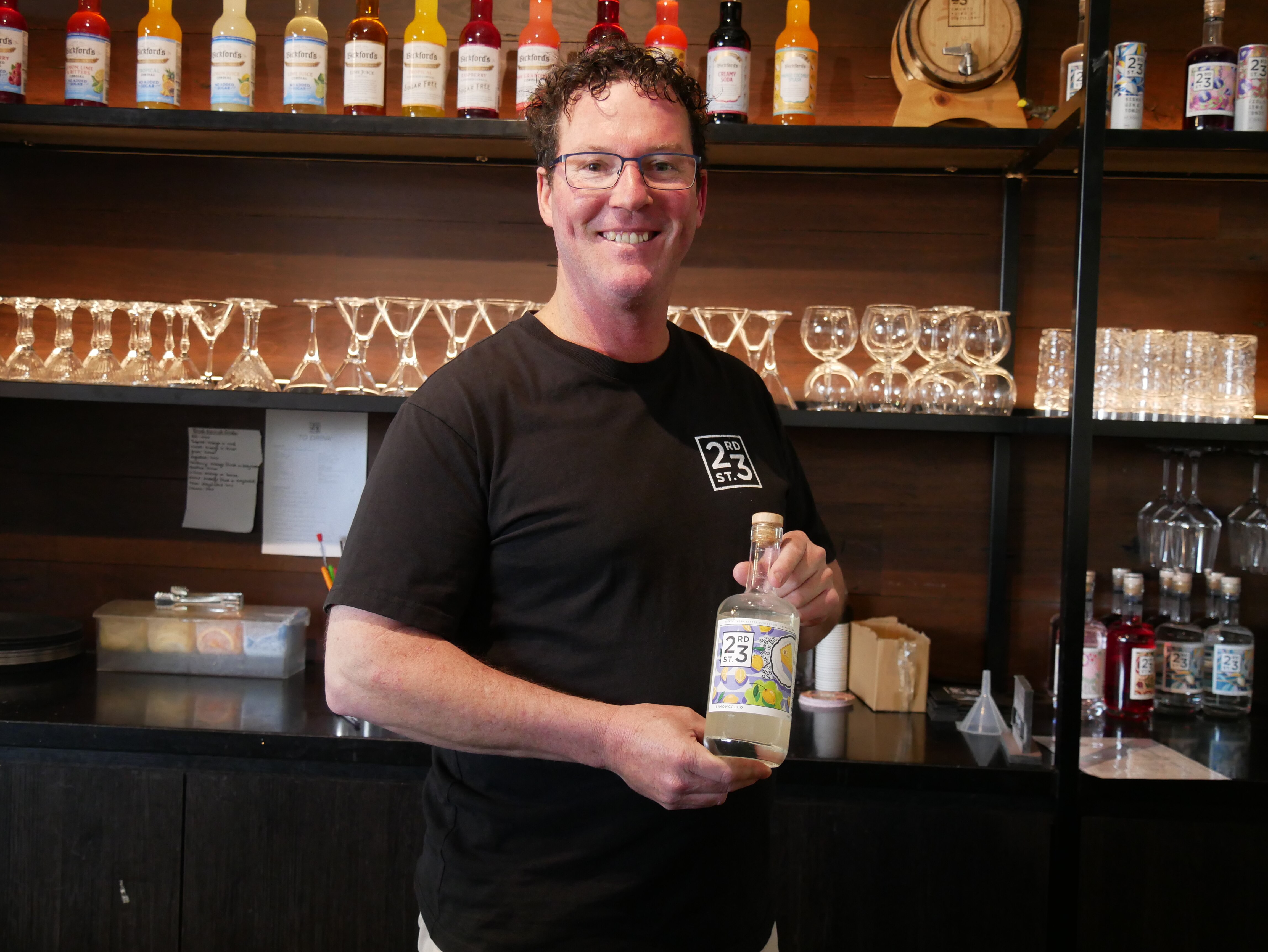 Un hombre de piel clara, gafas y cabello castaño rizado sonríe y sostiene una botella de limoncello detrás de una barra. 