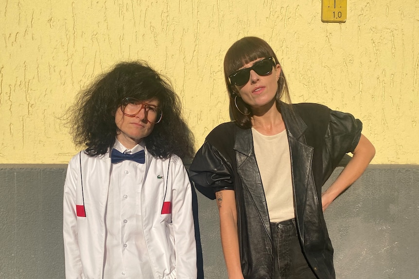 Two people stand against a wall. One is a person with long, bushy hair and glasses. The other is a woman with a fringe.