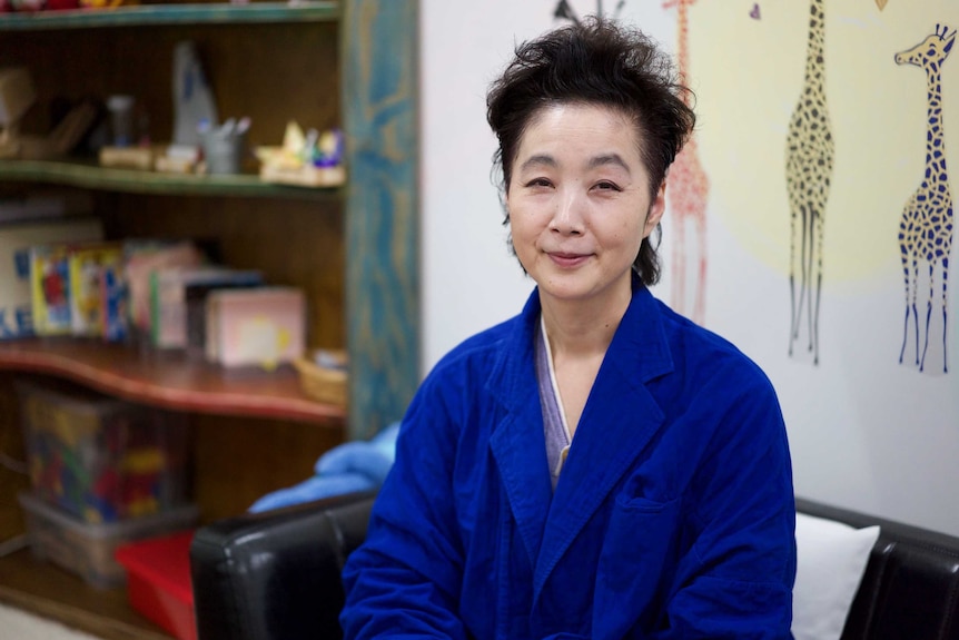 A Japanese woman in a lab coat in front of a children's mural