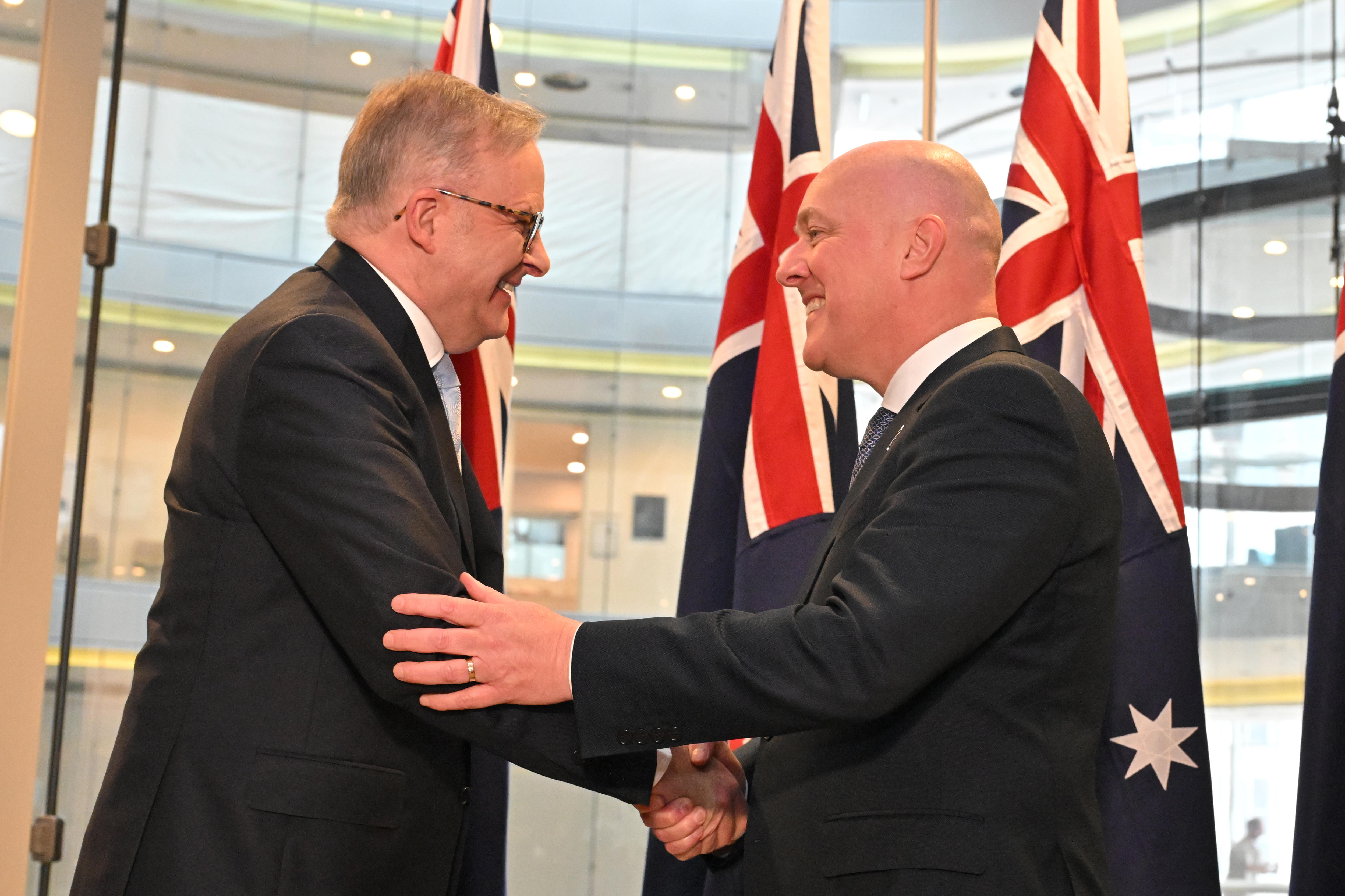 PM Anthony Albanese shakes hands with NZ PM Christopher Luxon in a warm greeting in front of flags from the two countries