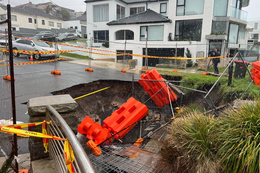 A sinkhole emerging in an affluent Sydney suburb, near the ocean, with rainclouds overhead.
