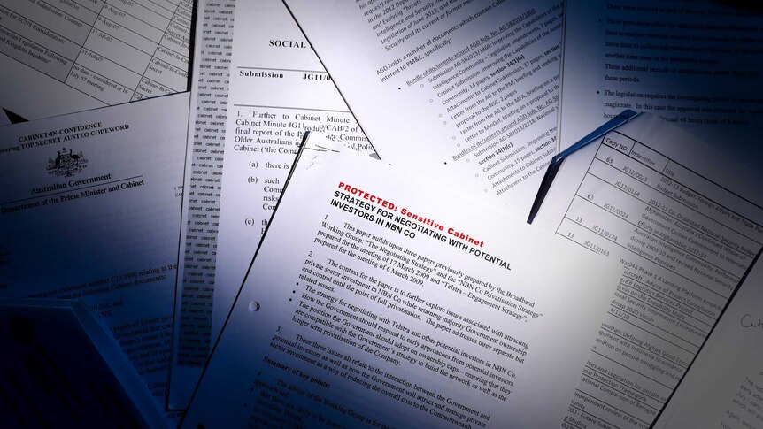 A pile of papers are scattered on a desk. A beam of light cuts across, with the top right and bottom left corners in shadow.