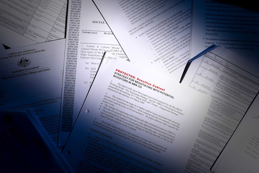 A pile of papers are scattered on a desk. A beam of light cuts across, with the top right and bottom left corners in shadow.